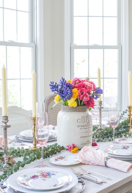 a spring tablescape with bright porcelain, a colorful floral centerpiece, a eucalyptus runner, candles and pink napkins