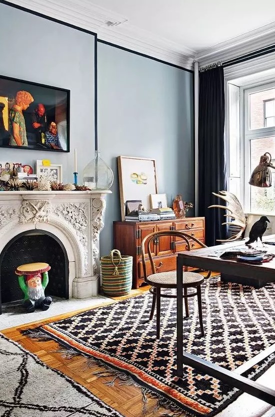 a boho eclectic home office with blue walls, an ornated fireplace, printed layered rugs, a dark desk and a rattan chair, a stained sideboard