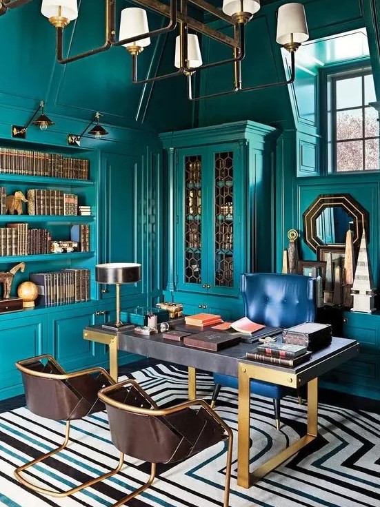 a bright maximalist home office with teal walls and matching built-in furniture and shelves, a refined desk, a navy and leather chairs, a chic chandelier