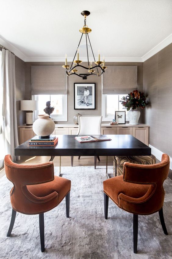 a catchy and chic taupe home office with a light stained console, a black desk, rust colored chairs and a chic chandelier