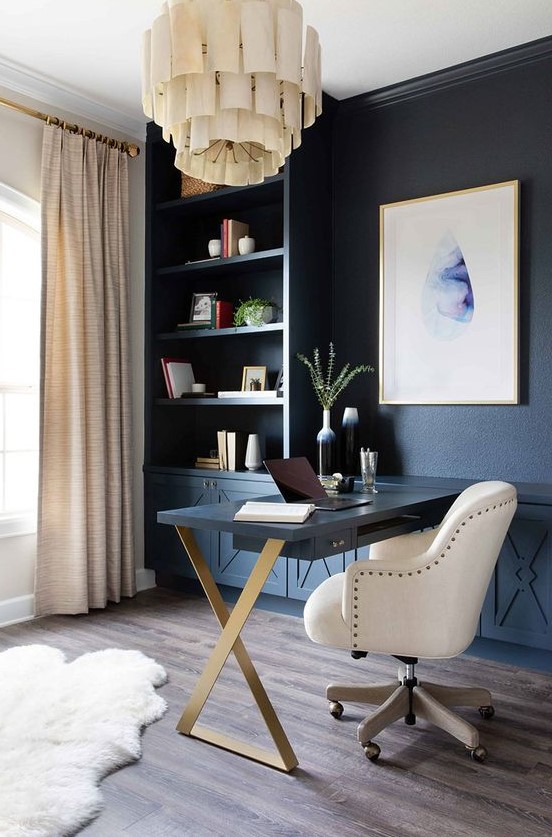 a chic home office with a blue wall taken by storage units, a statement artwork, a unique chandelier, a Murphy desk and a large white chair