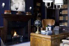 a chic moody home office with navy walls, built-in bookcases, a light-stained vintage desk, neutral chairs and a chic chandelier