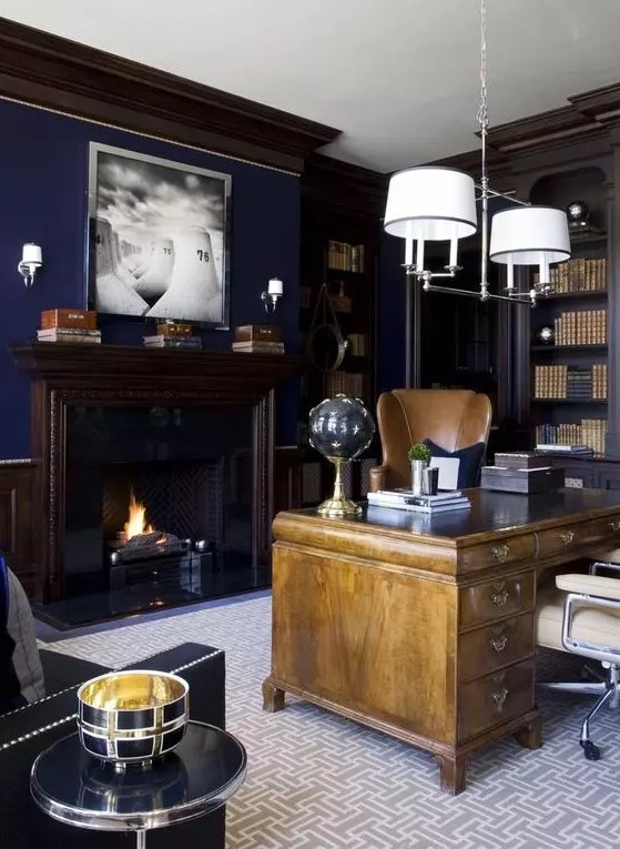a chic moody home office with navy walls, built in bookcases, a light stained vintage desk, neutral chairs and a chic chandelier