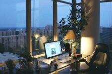 a contemporary home office with glazed wall, a black desk and a black leather chair, potted greenery and a tree and lights