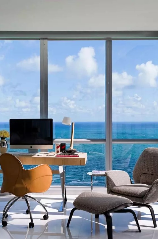 a fab home office with a glazed wall and a lovely sea view, a desk, an amber chair, a grey chair to relax in and a footrest