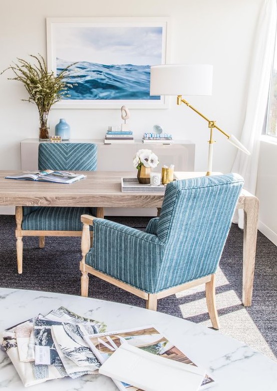 a gorgeous seaside home office with a wooden desk, blue chairs, a sea inspired artwork, an elegant and catchy table lamp