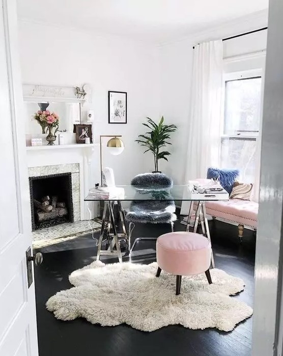 a light filled home office with a fireplace, a glass trestle desk, a pink sofa and a pouf, potted plants, brass lamps looks cute and cool