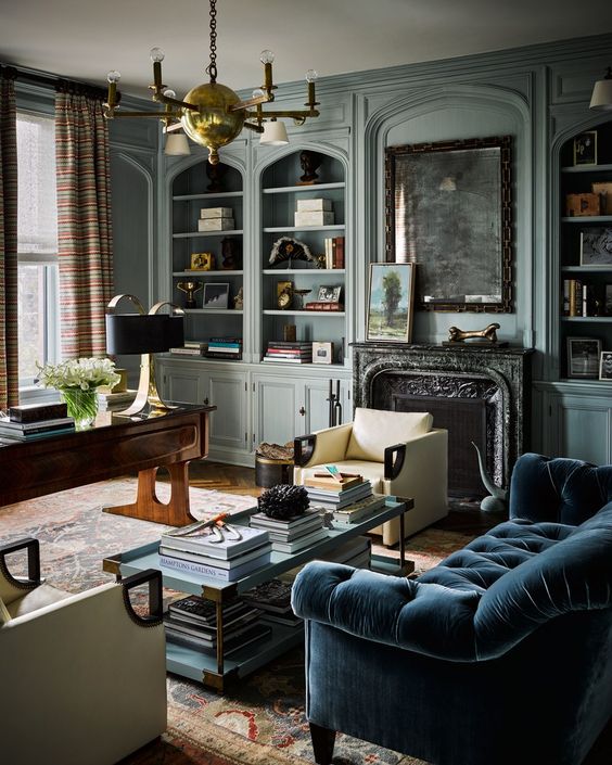 a lovely vintage home office with light blue walls, a fireplace, a dark-stained desk, a blue sofa, a tiered coffee table and a gilded chandelier