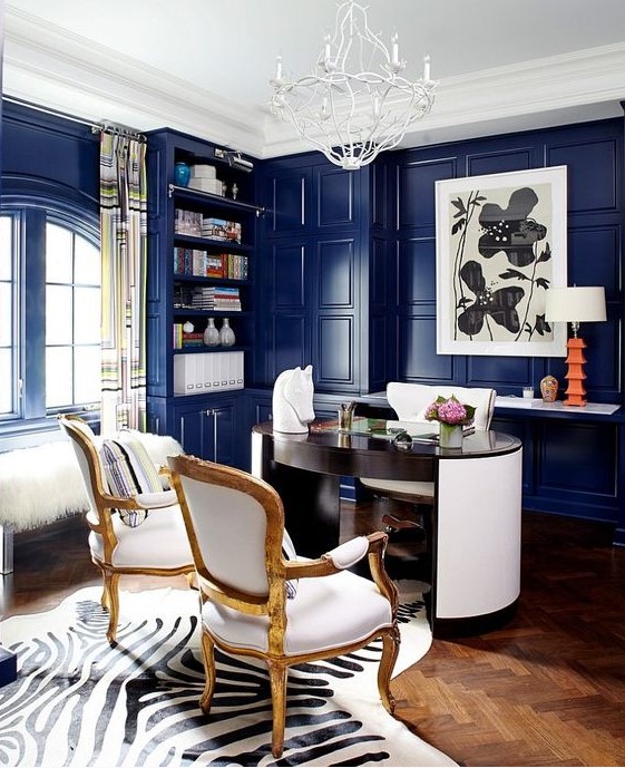 a luxurious blue home office with bright walls, a round desk, white furniture, a quirky chandelier, an acrylic bench with faux fur
