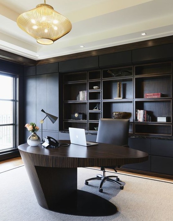 a minimalist and refined home office with a black built-in shelving unit, a sculptural oval desk of rich stained wood, a bold chandelier