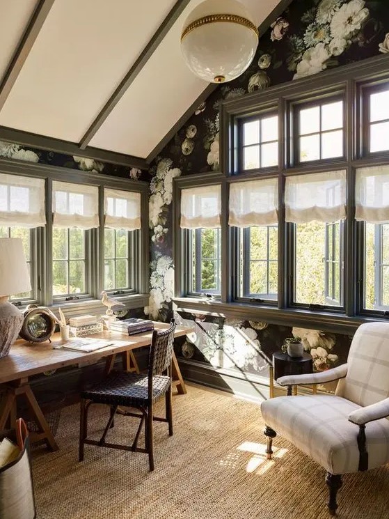 a modern country home office with moody floral wallpaper, a trestle desk, a woven leather chair, a plaid chair, a pendant lamp