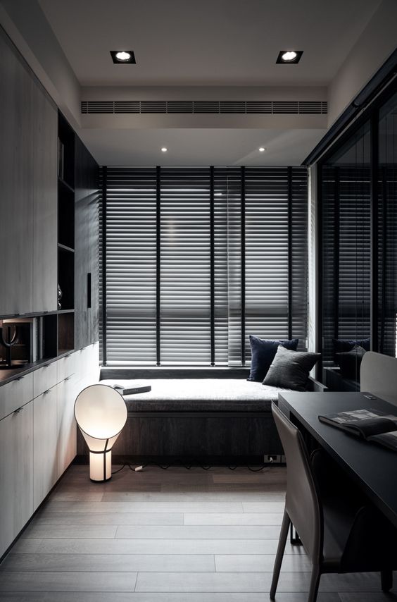 a moody contemporary home office with a windowsill daybed, a built-in storage unit, a black desk and black chairs and some lights