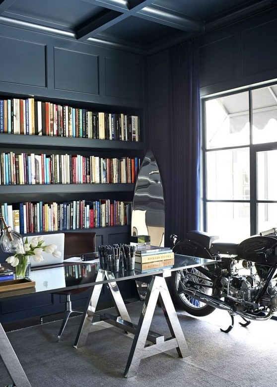 a navy home office with a coffered ceiling, built in storage space, a glass and metal desk plus a bike as decor