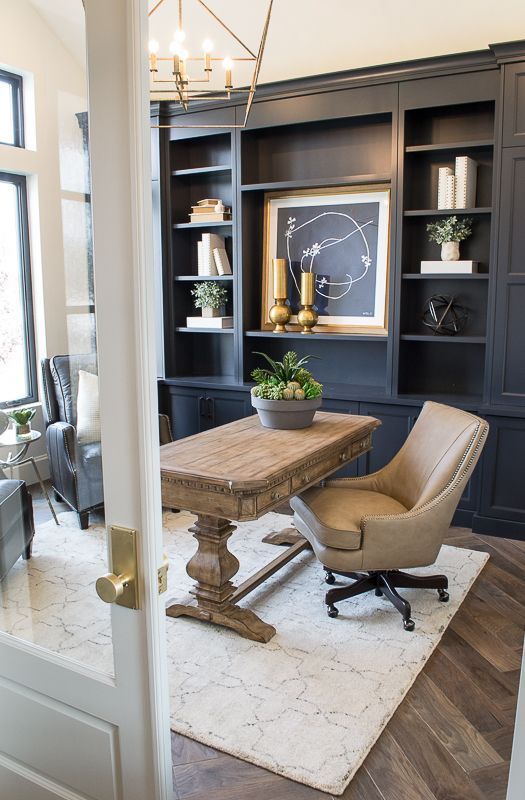 a pretty farmhouse home office with a grey storage unit, a stained rustic desk, a tan leather chair and artworks, a gilded chandelier
