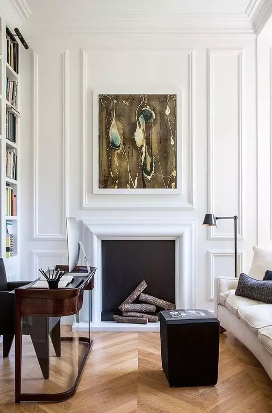 a sophisticated home office with white paneling, a non-working fireplace, a dark-stained rounded desk, a black pouf and a creamy sofa is wow