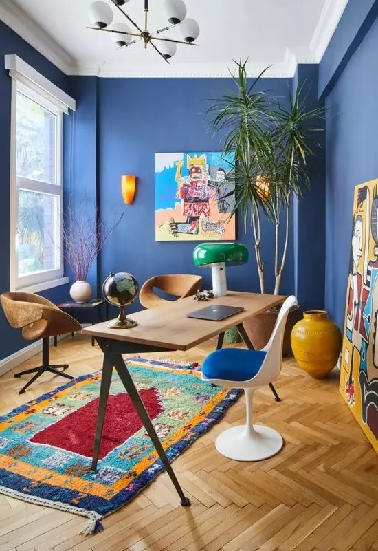 a vibrant home office with bold blue walls, colorful artworks and a rug, mismatching chairs and a statement plant in the corner