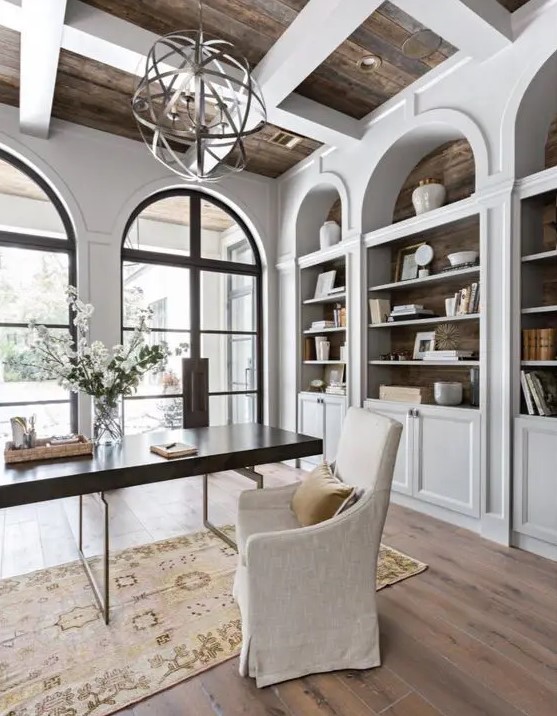 a vintage farmhouse home office with white walls and storage units, a sphere chandelier, a modern desk and an upholstered chair