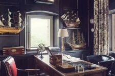 a vintage moody home office with black walls and cabinetry, a stained desk and a black and a red chair, ships for decor and a gold bubble lamp