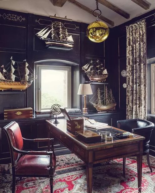 a vintage moody home office with black walls and cabinetry, a stained desk and a black and a red chair, ships for decor and a gold bubble lamp