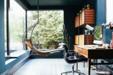 a welcoming home office in blue, with panoramic windows and a skylight, a wooden desk plus a leather chair, a hanging lounger and a shelving unit