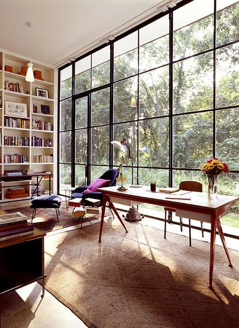 an airy and light-filled home office with a clear glass wall, built-in storage units, a desk and various chairs, cozy rugs