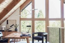 an attic home office with a wooden desk and a storage unit, black chairs, some lamps and a gorgeous view through double-height windows