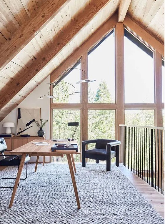 an attic home office with a wooden desk and a storage unit, black chairs, some lamps and a gorgeous view through double-height windows