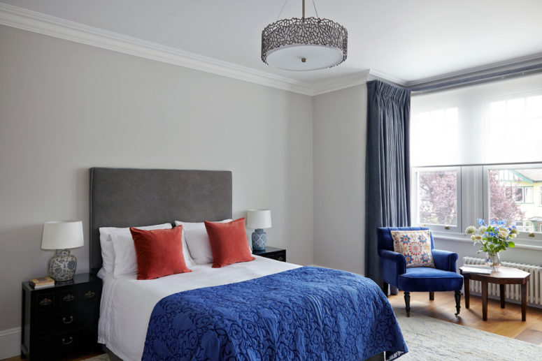 Master bedroom in period house with high ceiling and square bay window. Light grey walls brought to life with medium blue curtains, armchair and throw, contrasted with burnt orange cushions on the bed. Chinese style bedside tables with mismatched lamps add a quirky element. The armchair in the bay window offers a comfortable seat for reading a magazine when the opportunity presents.   