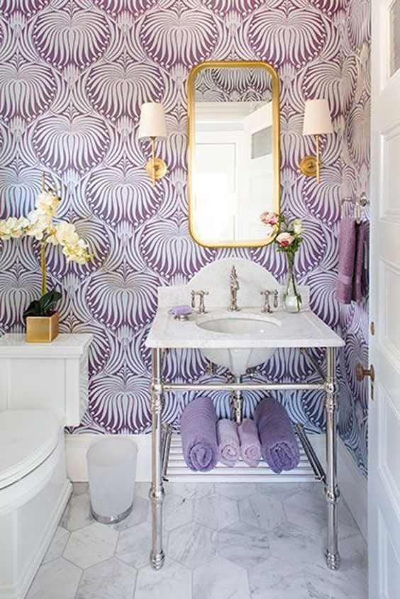 a beautiful purple and white bathroom with printed wallpaper, gilded touches and lamps and an orchid