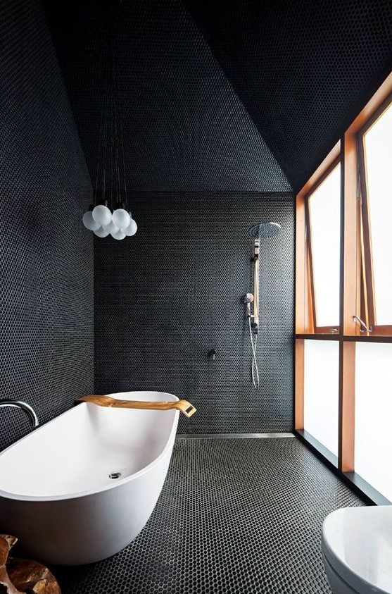 a black bathroom fully clad with penny tiles with white grout, with a frosted glass wall and a free standing bathtub