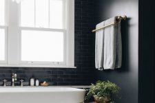 a black bathroom with black walls and tiles, a white tub, a white petal chandelier, a stained side table and greenery