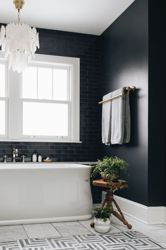 a black bathroom with black walls and tiles, a white tub, a white petal chandelier, a stained side table and greenery