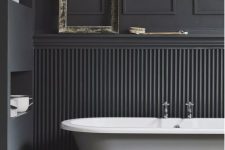 a black bathroom with paneling on the walls, niche shelves, a white clawfoot bathtub, a pendant lamp and a side table