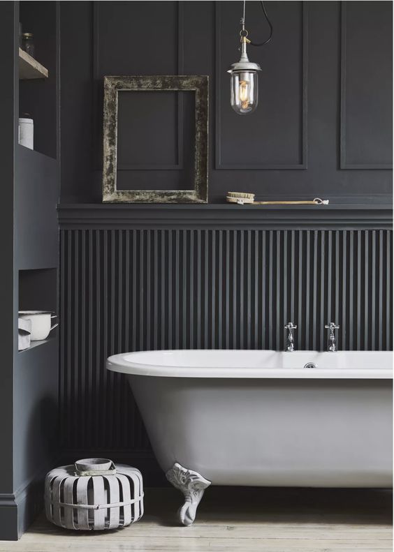 a black bathroom with paneling on the walls, niche shelves, a white clawfoot bathtub, a pendant lamp and a side table