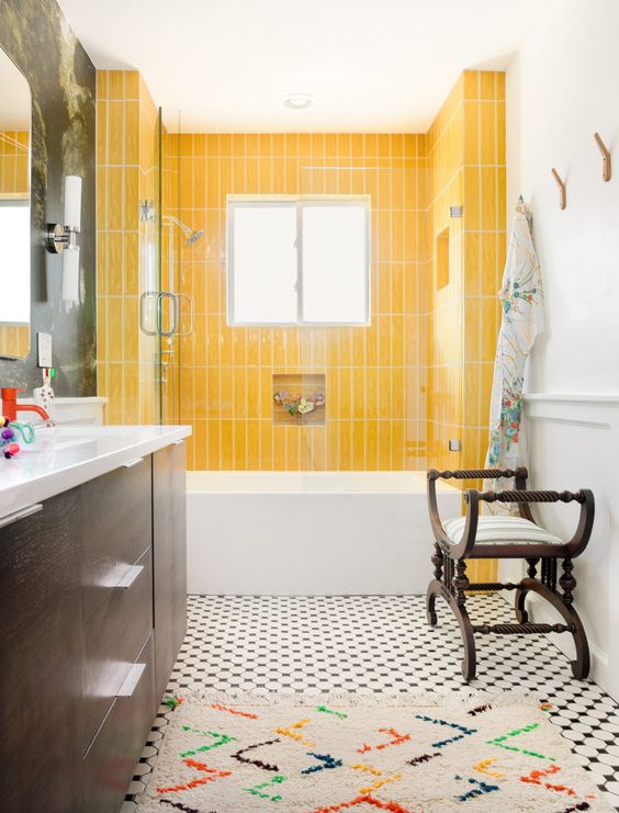 a bold bathroom with yellow skinny tiles, a printed tiled floor, a bright rug, vintage dark-stained furniture, floral towels is amazing