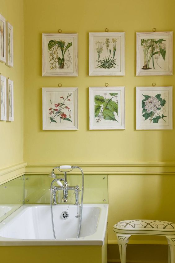 a bright yellow bathroom with a gallery wall of botanical posters, a bathtub with a glass backsplash and a vintage stool