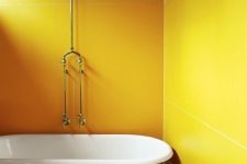 a bright yellow bathroom with super large yellow tiles, a yellow clawfoot bathtub, yellow tiles on the floor, a white bench and potted greenery