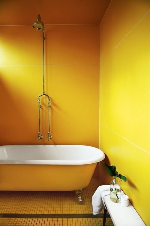 a bright yellow bathroom with super large yellow tiles, a yellow clawfoot bathtub, yellow tiles on the floor, a white bench and potted greenery