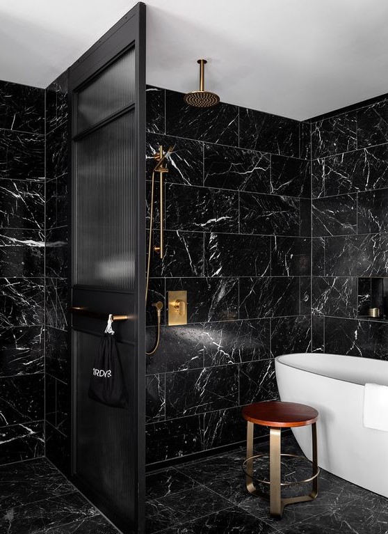 a chic black and gold bathroom with black marble tiles, black fixtures, a black space divider and a white tub