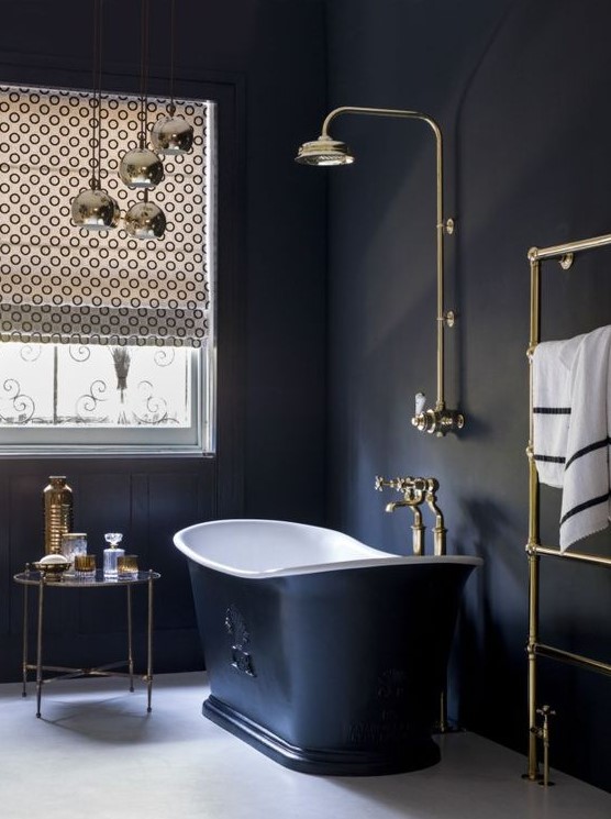 a chic vintage bathroom with black paneled walls, a black tub, gold fixtures, printed shades and gold accessories