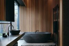 a contemporary black bathroom accented with light-stained wooden slabs, a black tub and black large scale tiles, a glazed wall and a stone platform
