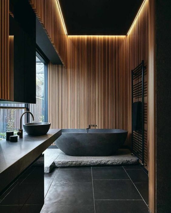 a contemporary black bathroom accented with light-stained wooden slabs, a black tub and black large scale tiles, a glazed wall and a stone platform