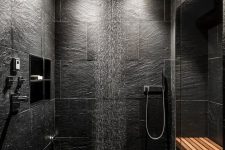 a contemporary black bathroom done with textural black tiles, a rain shower and a wooden bench
