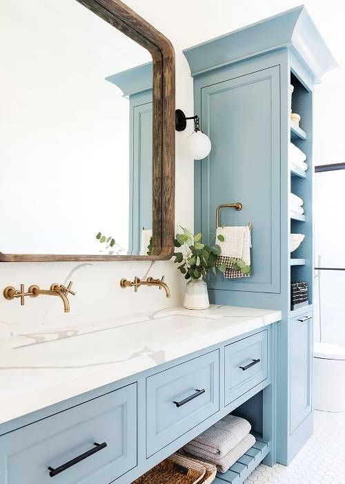 a cozy rustic bathroom with light blue furniture, a wooden frame mirror and brass fixtures