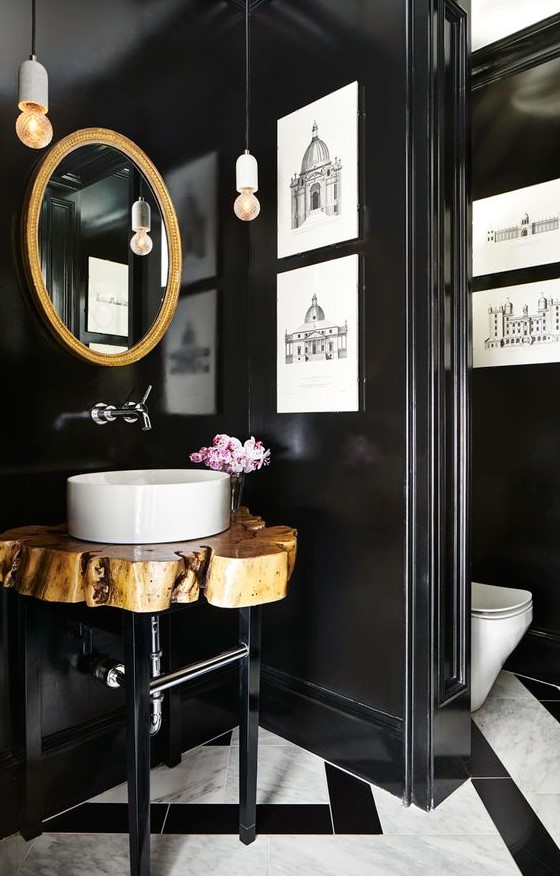 a gorgeous bathroom with a mosaic tile floor, black walls, a gold frame mirror, a wood slice vanity and artworks