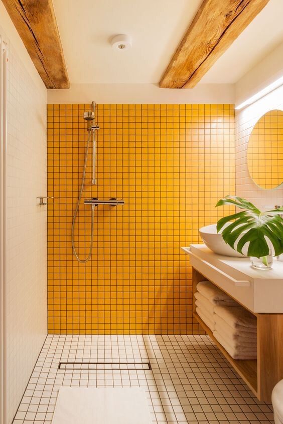 a laconic modern bathroom clad with yellow and neutral square tiles, a floating vanity with a bowl sink, wooden beams and a round mirror