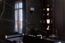a luxurious black bathroom with a marble accent wall and a bathtub clad with it, too, corner shelves with decor and a gold knot chandelier