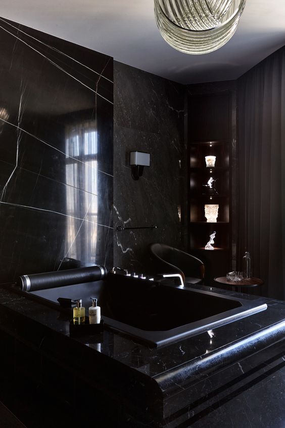 a luxurious black bathroom with a marble accent wall and a bathtub clad with it, too, corner shelves with decor and a gold knot chandelier