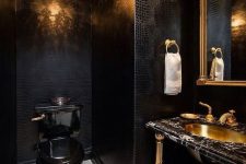 a luxurious powder room with black leather walls, a crystal chandelier, a black toilet, a black marble vanity and a gold sink