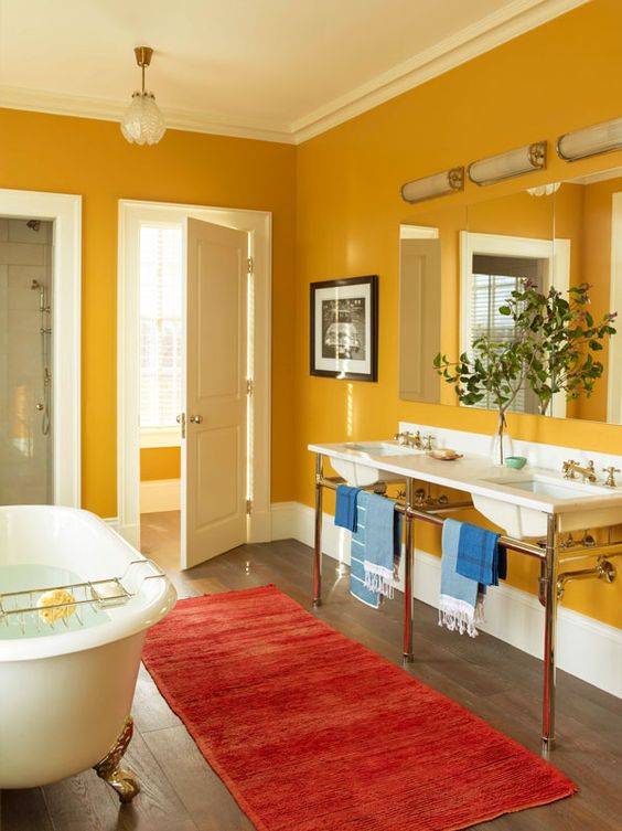 a modern bathroom with warm yellow to mustard walls, sinks on pretty stands, a large mirror with lights, an oval tub and a bold red rug